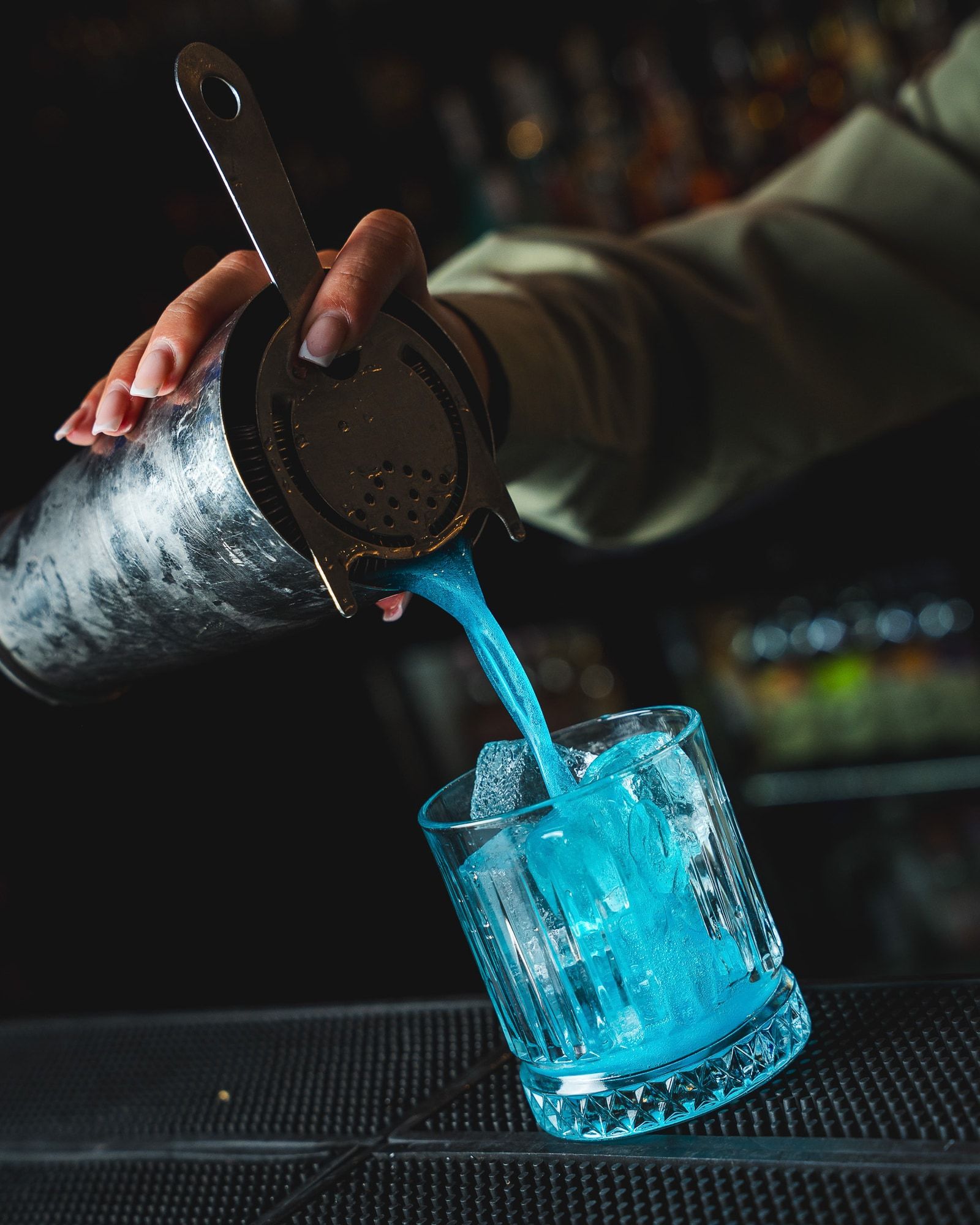 A blue cocktail being poured from a shaker into a glass of ice.